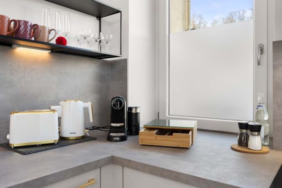 Moderne Küche mit Wasserkocher, Kaffeemaschine und Holzregal. Tageslicht fällt durch das Fenster.