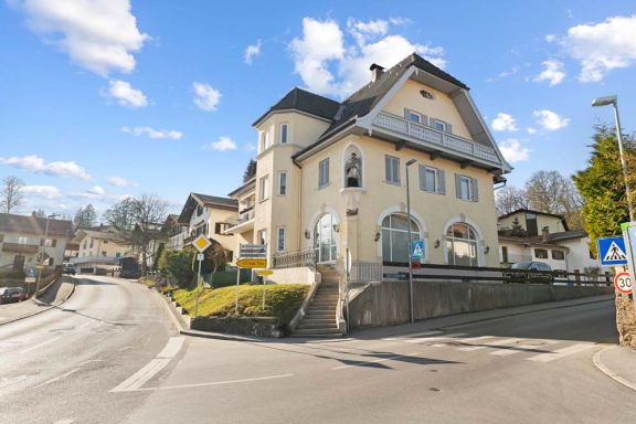 Ein helles, freistehendes Gebäude mit Rundbogenfenstern und umgebenden Bäumen.