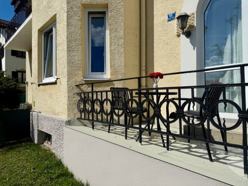 Ein Balkon mit schwarzem Geländer und zwei Stühlen, daneben ein rotes Objekt.