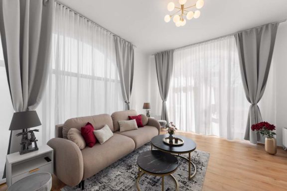 Helles Wohnzimmer mit beiger Couch, runden Tischen und großen Fenstern.